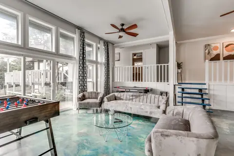 Turquoise-floored game lounge with foosball table, air hockey table, plush gray channel-tufted seating, and glass coffee table beside tall windows at Brompton Manor, Oklahoma City
