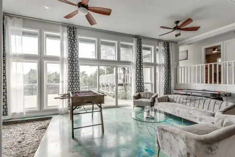Sunlit lounge with floor-to-ceiling windows, gray velvet sofas, foosball table, and air hockey table on a turquoise stained-concrete floor at Brompton Manor, Oklahoma City