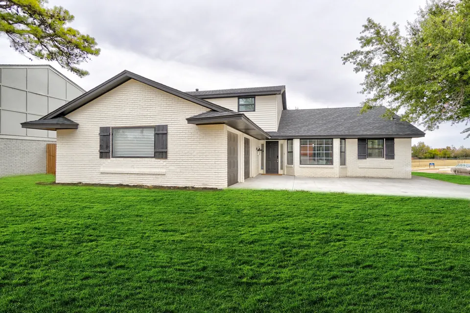 White-painted brick home with black shutters, attached garage, and a wide green front lawn at The Village Manor, Oklahoma City