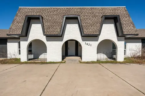 White-painted brick front exterior with three dramatic arched porch openings, mansard-style shingle roof, and wide concrete driveway under a blue sky at Will Rogers Manor, Oklahoma City